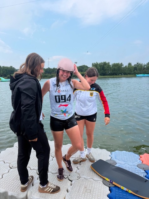 Kyra Hartan mit Startnummer 094 und rosafarbenem Helm, nass und lachend auf dem Ponton am Wasser, wird nach ihrem Finallauf von zwei jungen Frauen in Empfang genommen – eine davon in der Jacke des deutschen Teams. Rechts im Bild liegt ihr Wakeskate.