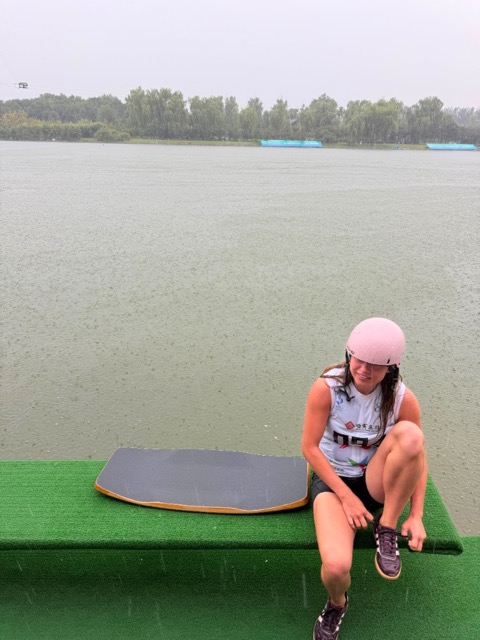Kyra Hartan im Regen am Ufer des Shunyi Olympic Cable Park, auf einer Kunstrasenbank sitzend und ihre Schuhe bindend, neben sich ihr Wakeskate.
