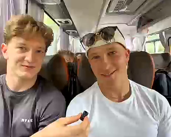 Carlo Hüsing im grauen Longsleeve und Max Bamler im weißen Shirt nebeneinander auf der Rückbank des Teambusses, Carlo hält ein Ansteckmikrofon.